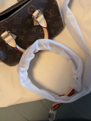 Louis Vuitton monogram handväska - Säljer en klassisk Louis Vuitton handväska i brunt monogrammönster med beige och röda detaljer. Väskan har två handtag och avtagbar axelrem, samt guldfärgade metalldetaljer. Kommer med original dustbag och box.