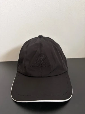 Svart keps  - Snygg svart keps från med diskret broderad logga framtill. Kepsen har böjd skärm med vit kant och klassisk sexpanelsdesign. Insidan är fodrad med rutigt tyg och har en brun kant. Perfekt accessoar för en stilren look.