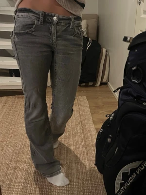 Grå bootcut jeans med låg midja - Säljer ett par grå bootcut jeans med låg midja och klassiska fem fickor. Jeansen har en lätt tvättad look och raka ben som går ut lite nedtill. Perfekta för en avslappnad och trendig stil.
