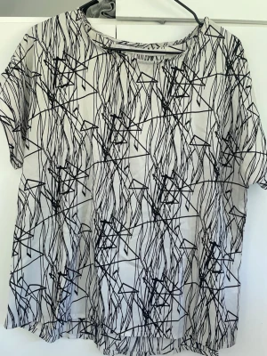 Vit t-shirt med svart abstrakt mönster - Säljer en vit t-shirt med svart abstrakt mönster. T-shirten har korta ärmar och en avslappnad passform. Perfekt för dig som gillar grafiska prints och vill sticka ut lite extra.