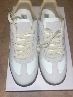 Maison Margiela vita sneakers - Maison Margiela sneakers i vitt med beige detaljer och gummisula. Ovandelen är i en mix av mocka och slätt läder, med klassiska snörningar och låg profil. Snygg minimalistisk design som passar till det mesta.
