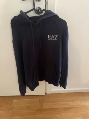  EA7 Emporio Armani hoodie - Snygg mörkblå hoodie från EA7 Emporio Armani med vit logga på bröstet och tryck på ärmen. Hoodien har en stor huva med dragsko och en klassisk magficka framtill. Perfekt för en avslappnad och trendig stil.