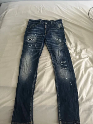 Mörkblå slitna jeans med färgstänk - Säljer ett par mörkblå jeans med slitningar och vita färgstänk för en cool, avslappnad look. Jeansen har raka ben, klassisk femficksmodell och detaljerade slitningar på både fram- och baksida. Perfekta för dig som gillar streetstyle och unik design.