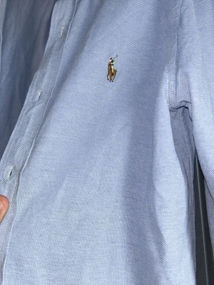 Ljusblå skjorta från Ralph Lauren - Klassisk ljusblå skjorta från Ralph Lauren med broderad logga på bröstet. Skjortan har knappar framtill och lång ärm, tillverkad i mjuk bomull. Perfekt för en clean och stilren look.