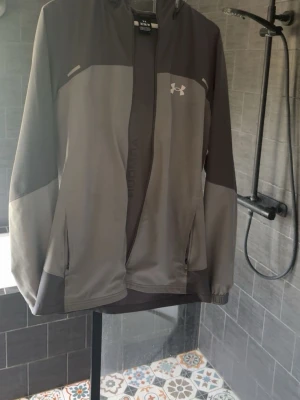Grå vindjacka från Under Armour - Snygg grå vindjacka från Under Armour med dragkedja framtill och två sidofickor. Jackan har huva med dragsko och logga på bröstet. Tillverkad i lätt och vindtätt material, perfekt för blåsiga dagar.