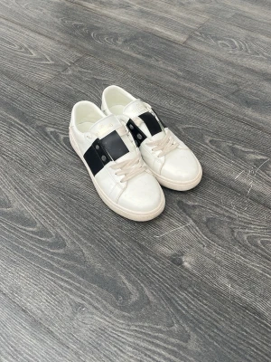 Vita sneakers med svart detalj - Snygga vita sneakers med bred svart rem över vristen och vita snören. Skorna har en klassisk låg profil och en stilren design som passar till det mesta. Tillverkade i slätt material med gummisula och metallnitar på sidan av remmen.