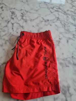 Röda badbyxor från Sail Racing - Snygga röda badbyxor från Sail Racing med svart dragsko i midjan och logga längs benet. Klassisk modell med elastisk midja och snabbtorkande material, perfekta för stranden eller poolen.