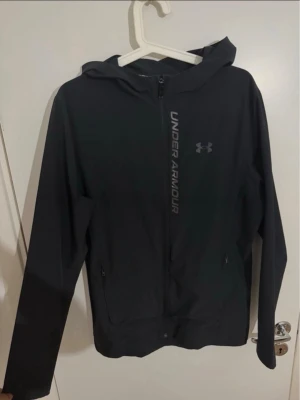 Svart vindjacka från Under Armour - Snygg svart vindjacka från Under Armour med huva och dragkedja framtill. Jackan har två sidofickor med dragkedja och logga samt text på bröstet. Tillverkad i lätt och funktionellt material som andas.