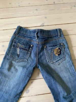 Blå bootcut jeans med broderi - Säljer ett par lågmidjade blå bootcut-jeans med snygga slitningar och broderade detaljer på bakfickorna. Jag skulle säga att de känns som XS eller XXS.   Midjemåttet är 36 cm. Säljer då de är för små för mig. Det är bara att skriva om ni vill ha fler mått eller bilder! Är öppen för prisförslag💕