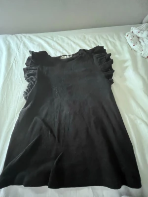 Svart ärmlös topp med volang - Snygg svart topp med volangdetaljer på axlarna. Toppen är ärmlös och har en rund halsringning. Perfekt för dig som vill ha en enkel men ändå trendig look. Materialet känns mjukt och följsamt.