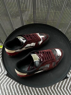 Valentino rockrunners  - Snygga sneakers från Valentino med vinröda mockadetaljer och kamouflagemönster i svart, vitt och grått. Skorna har svarta snören, svart innersula och beige hälparti. Perfekta för dig som vill sticka ut med en unik och trendig look. Priset är ej hugget i sten!
