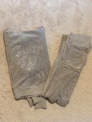 Grått Juicy Couture set med strass - Säljer ett superfint grått mjukiset från juicy couture i velour med glittrande stenar. Mycket bra kvalité, knappt använt och inga stenar saknas. Storlek S, midjemått koftan: 41cm, armlängd från armhålan: 48cm. Midjemått byxor: 35cm, innerbenslängd: 76cm.                             Priset är förhandlingsbart! Kontakta för fler bilder/frågor💕