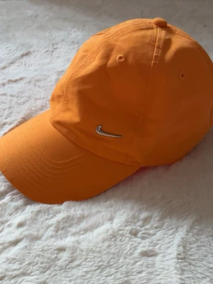 Orange keps från Nike - Snygg orange keps från Nike med klassisk böjd skärm och silverfärgad Swoosh-logga på framsidan. Tillverkad i lätt och bekvämt material, perfekt för en sportig och avslappnad stil.