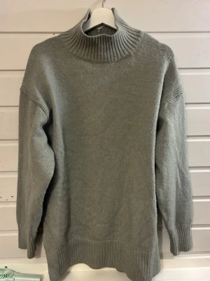 Ljusgrå stickad polotröja - Mysig ljusgrå stickad tröja med hög ribbad polokrage och lång ärm. Tröjan har en slits bak i kragen för extra detalj och är rak i modellen. Perfekt för kalla dagar och enkel att matcha till olika outfits.