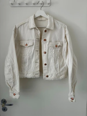 Vit jeansjacka från H&M - Snygg vit jeansjacka från H&M med klassisk krage, bröstfickor med lock och knappar i kopparfärg. Jackan har rå kant nertill och en avslappnad passform som ger en cool vibe. Perfekt att slänga över en hoodie eller t-shirt.