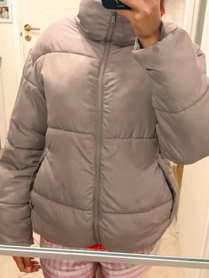 Ljusgrå pufferjacka med hög krage - Säljer en ljusgrå pufferjacka med hög krage och dragkedja framtill. Jackan har en oversized passform och breda kanaler som ger en riktigt fluffig look. Perfekt för kalla dagar och riktigt skön att bära. Materialet är slätt och ger en modern vibe. Den har några fläckar och priset kan diskuteras 
