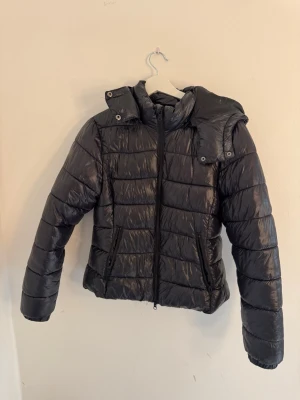 Svart pufferjacka med huva - Snygg svart pufferjacka med glansig finish och vadderad design. Jackan har dragkedja framtill, två fickor med dragkedja och en stor, mysig huva. Perfekt för kalla vinterdagar och ger en modern look.