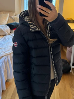 Svart dunjacka COLMAR  - Snygg svart dunjacka med dragkedja och huva. Jackan har ett unikt mönstrat innerfoder i svartvitt med blixtar och en patch på ärmen. Puffig modell som håller värmen under kalla dagar. Perfekt för dig som vill ha en stilren men ändå cool vinterjacka.