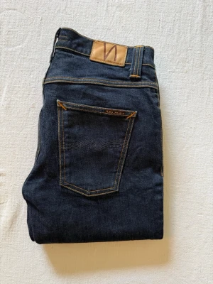 Mörkblå jeans från Nudie Jeans - Snygga mörkblå jeans från Nudie Jeans med klassisk femficksmodell och kontrastsömmar. Jeansen har rak passform och är tillverkade i kraftig denim. Perfekta för dig som gillar stilrena och tidlösa jeans. Storlek W32 L34!