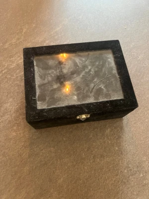 Svart smyckeslåda med glaslock - Snygg svart smyckeslåda med glaslock och mjuk sammetsinsida. Perfekt för att förvara ringar, örhängen eller andra småsmycken. Lådan har flera fack och en praktisk låsanordning framtill. Stilren och enkel design som passar alla.