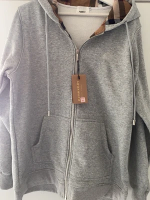 Grå zip hoodie från Burberry - Snygg grå hoodie med dragkedja från Burberry. Klassisk rutmönstrad insida på huvan i beige, brunt och vinrött. Mjuk fleece på insidan, stora fickor fram och justerbar huva med snören. Perfekt för en chill och stilren look.