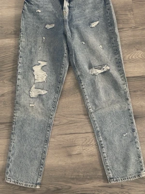 Grå mom jeans med slitningar - Säljer ett par grå mom jeans med flera slitningar och hål på benen. Jeansen har hög midja, raka ben och klassisk femficksmodell. Materialet är kraftig denim i bomull och tvätten är ljusgrå. Perfekta för en avslappnad och trendig look. 38 storlek, 32 längd 