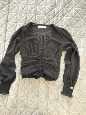 Svart blus med knappar och v-ringning - Snygg svart blus från Chiara Forti med v-ringning, klädda knappar framtill och smockade muddar vid ärmsluten. Blusen har ett figurnära snitt med rynkade detaljer i midjan och volangkant nedtill. Tillverkad i ett glansigt, tunt material som ger en elegant look.