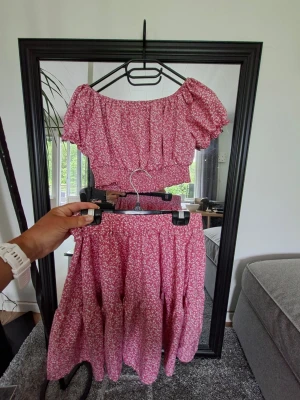 Rosa blommigt tvådelat set - Säljer ett sött tvådelat set med en kort topp och en vid kjol i rosa med små vita blommor. Toppen har puffärmar och resår i midjan, kjolen har elastisk midja och volanger. Perfekt för dig som gillar romantisk och somrig stil.
