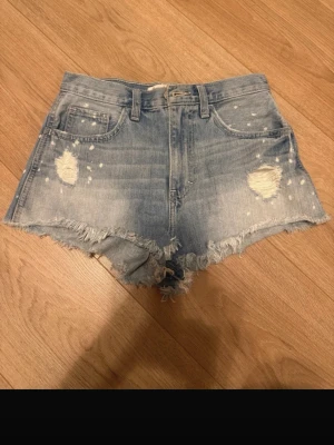 Ljusblåa jeansshorts Hollister - Säljer ett par ljusblåa högmidjade jeansshorts med slitna detaljer och fransig kant. Shortsen har klassisk femficksdesign, dragkedja och knapp framtill samt coola slitningar och vita stänk för en trendig look.