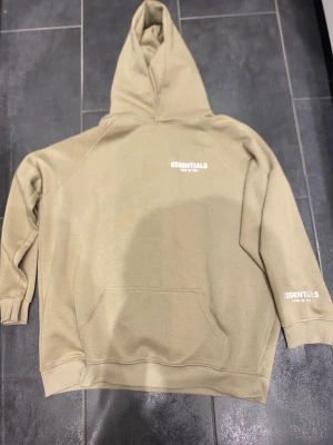 Beige Essentials Fear of God hoodie - Snygg beige hoodie från Essentials Fear of God med vit logga på bröstet och ärmen. Klassisk känguruficka framtill och stor huva. Tillverkad i mjukt bomullsmaterial som känns skönt mot huden. Perfekt för en avslappnad streetwear-look.