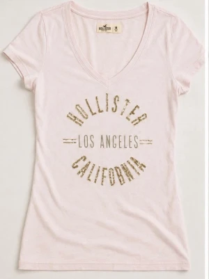 Ljusrosa Hollister t-shirt v-ringad - Snygg ljusrosa t-shirt från Hollister med v-ringning och tryck i beige/guld med texten 'Hollister Los Angeles California' på bröstet. Kortärmad och i mjuk bomull som sitter skönt och har en slim passform.