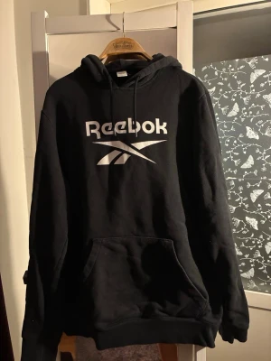 Svart hoodie från Reebok med logga - Säljer en svart hoodie från Reebok med stor vit logga på bröstet. Hoodien har huva med dragsko och en stor magficka framtill. Tillverkad i mjukt sweatshirtmaterial som är perfekt för en avslappnad stil.