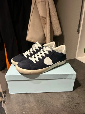 Mörkblå sneakers från Philippe Model - Snygga mörkblå sneakers från Philippe Model med vita detaljer och vit sula. Skorna har låga skaft, vita skosnören och en ikonisk logotyp på sidan. Ovandelen är i mocka och läder vilket ger en exklusiv känsla. Perfekta för dig som gillar stilrena och trendiga sneakers.