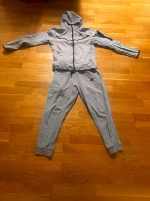 Grå zip-hoodie från Nike - Grå zip-hoodie från Nike med huva och dragkedja framtill. Tröjan har långa ärmar, ribbade muddar och en liten Nike-logga på bröstet. Perfekt för en avslappnad och sportig stil. Med byxor alltså en Nike tech tracksuit.