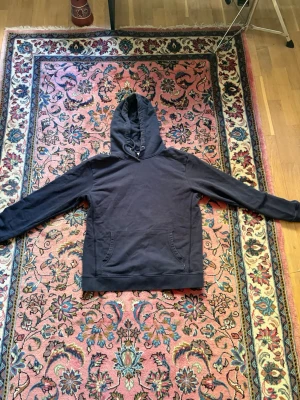 Mörk hoodie från Les Deux - Säljer en mörk hoodie från Les Deux med broderad logga på bröstet. Hoodien har huva med dragsko och en klassisk magficka. Tillverkad i mjukt bomullsmaterial och har ribbade muddar vid ärmslut och nederkant. Perfekt för en chill och stilren look.