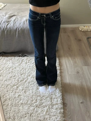 Mörkblå bootcut jeans med kontrastsömmar - Säljer ett par mörkblå bootcut jeans med vita kontrastsömmar och låg midja. Jeansen har klassiska fem fickor och markerade sömmar både fram och bak. Materialet är denim med lätt stretch för skön passform. 
