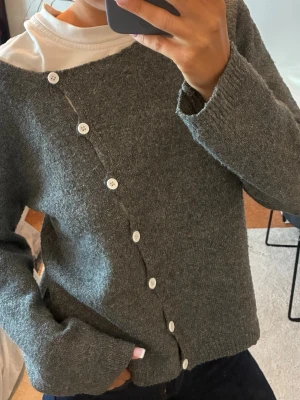 Grå asymmetrisk kofta med knappar - Säljer en grå kofta med unik asymmetrisk knäppning och vita knappar. Koftan har lång ärm och är stickad i ett mjukt material. Perfekt för dig som gillar en stilren men ändå lite annorlunda look.