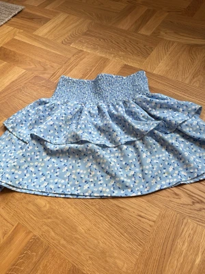 Blommig blå volangkjol - Söt blå kortkjol med vita och blå blommor, bred smock i midjan och dubbla volanger. Kjolen har en luftig och lätt känsla, perfekt för varma dagar. Passar dig som gillar en feminin och somrig stil.