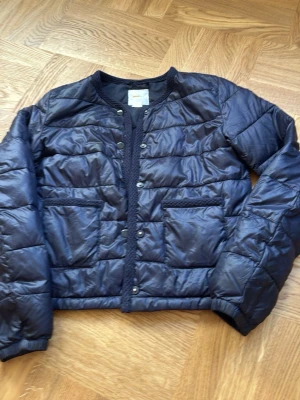 Mörkblå pufferjacka från Gina Tricot - Snygg mörkblå pufferjacka från Gina Tricot med rund halsringning och tryckknappar framtill. Jackan har två stora fickor fram och quiltad design. Materialet är glansigt och ger en modern look, perfekt för kalla dagar.