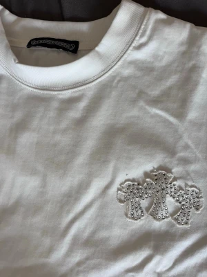 Vit t-shirt med broderade kors - Vit t-shirt i bomull med broderade kors och små silverfärgade nitar på bröstet och ryggen. Klassisk rund hals och korta ärmar. Snygg detalj med broderad text på ryggen. Perfekt för dig som gillar stilrena plagg med edge.