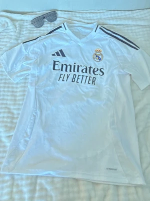 Vit Real Madrid matchtröja Adidas - Säljer en vit Real Madrid matchtröja från Adidas med klubbmärke på bröstet och svarta detaljer på axlarna. Tröjan har trycket 'Emirates Fly Better' på framsidan och är tillverkad i ett lätt, ventilerande material perfekt för fotboll eller träning.