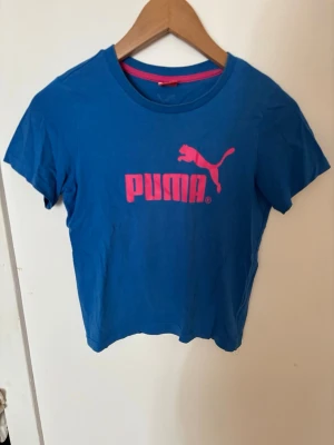 Blå Puma t-shirt med rosa logga - Snygg blå t-shirt från Puma med korta ärmar och rosa logga framtill. T-shirten har en klassisk rund halsringning och är tillverkad i mjuk bomull💙