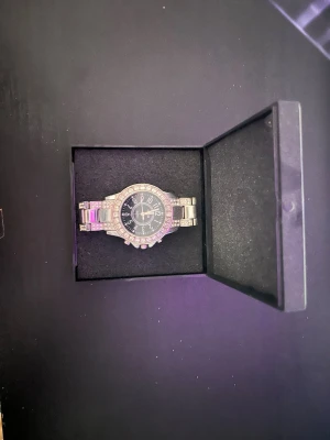 Silverfärgad klocka med stenar - Stilren silverfärgad klocka med metallarmband och glittrande stenar runt urtavlan. Urtavlan är svart med tydliga siffror och detaljer i silver. Klockan har en klassisk rund form och känns både lyxig och trendig.