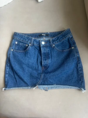 Blå jeanskjol med rå kant - Säljer en klassisk blå jeanskjol från Bik Bok med  fickor och rå, fransig kant nedtill. Kjolen har normal passform och är i ett slitstarkt denimtyg. Perfekt till sommaren för en avslappnad look.