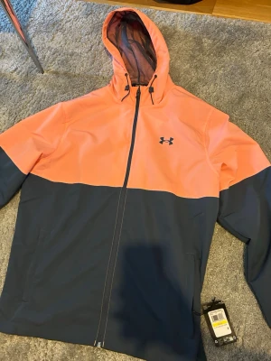 Under Armour vindjacka orange/marin - Snygg vindjacka från Under Armour i tvåfärgad design med orange upptill och marinblå nedtill. Jackan har huva med dragsko, hel dragkedja och logga på bröstet. Perfekt för blåsiga dagar och sportiga outfits.