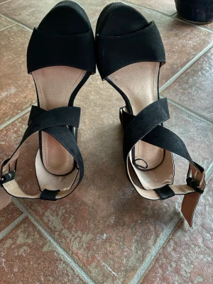 Svarta pumps med bred klack - Snygga svarta pumps med bred klack och öppen tå. Skorna har korsade remmar över foten och justerbart spänne vid ankeln. Materialet ser ut att vara mockaimitation och innersulan är ljus. Perfekta för dig som gillar stilrena och bekväma klackar. Kolla gärna mina andra annonser❗️