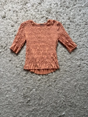 Rostfärgad smockad topp med volang - Supersnygg orange topp med smock över hela kroppen och ärmarna. Toppen har trekvartsärm och en söt volangkant nedtill. Perfekt för dig som gillar en unik och trendig look.