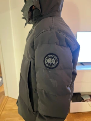 Svart pufferjacka från Canada Goose - Säljer en svart/grön pufferjacka från Canada Goose med huva och diskret logotyp på ärmen. Jackan har raka linjer, stora fickor och stängs med knappar och dragkedja. Perfekt för kalla vinterdagar och riktigt varm tack vare tjock vaddering.