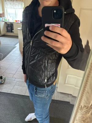 Svart John ricci cardigan - Snygg svart vindjacka med dragkedja framtill och två fickor med dragkedja. Jackan har en quiltad framsida och mjuka ärmar samt huva i sweatshirtmaterial. Perfekt för dig som vill ha en sportig och stilren look.