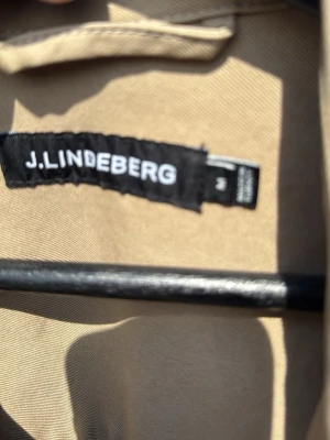 Beige overshirt från J.Lindeberg - Stilren beige overshirt från J.Lindeberg med två bröstfickor med lock och knappar. Klassisk krage och knappstängning framtill. Tillverkad i ett mjukt, slitstarkt tyg som passar perfekt för lager-på-lager-stil.